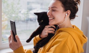 A woman in a golden yellow hoodie holds a black cat while using her smartphone.