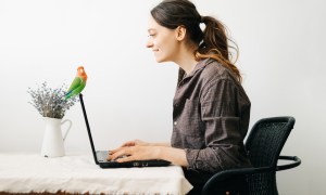 Woman sits at laptop while her pet lovebird perches on it