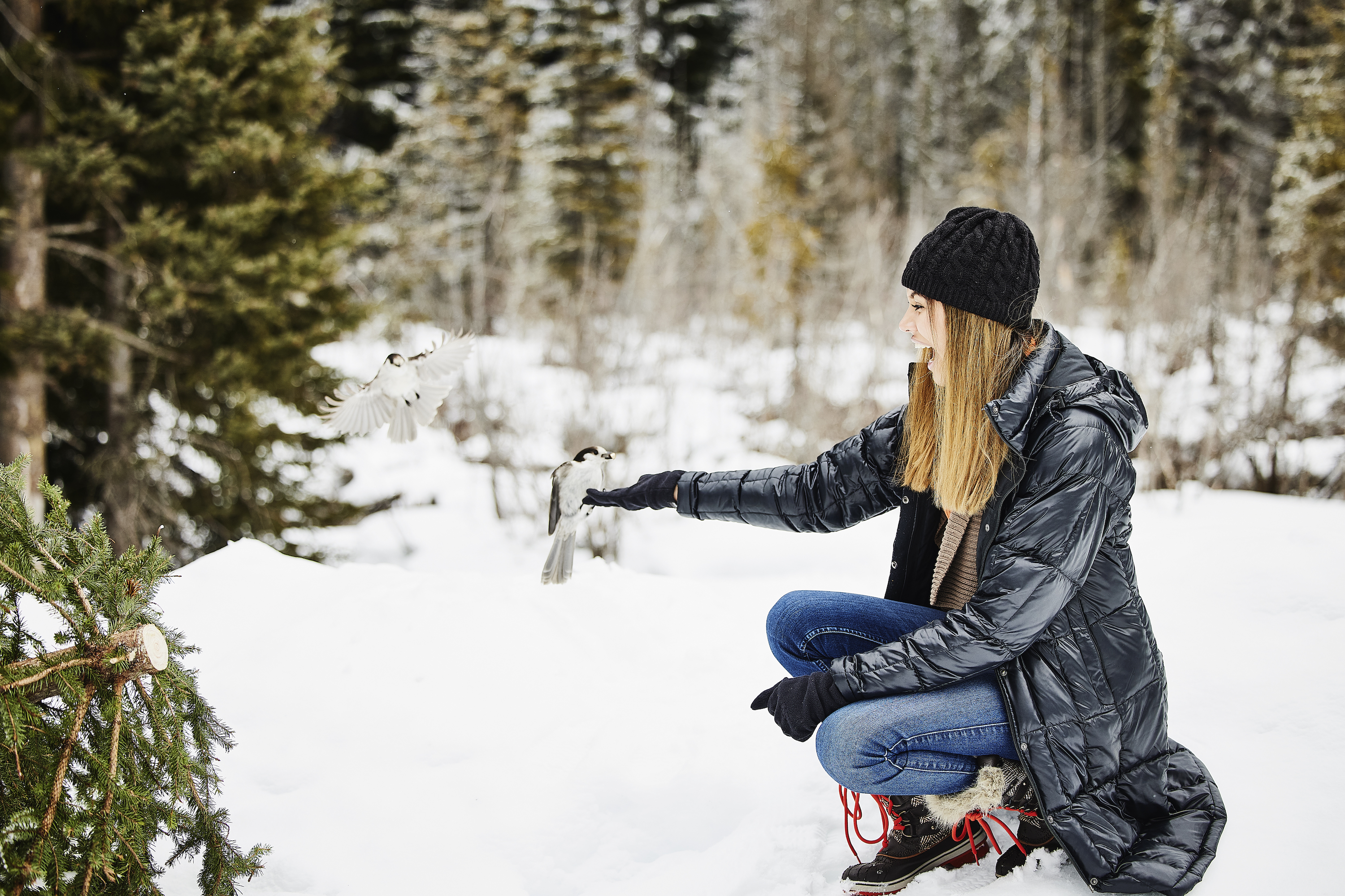 Wide shot of smiling young woman feeding bird out of hand in snowy field on winter afternoon